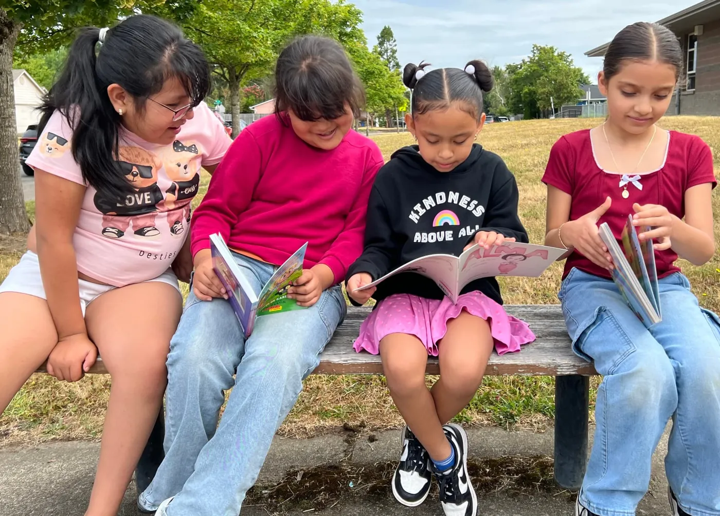 Four girls sit on a bench outdoors reading their new books